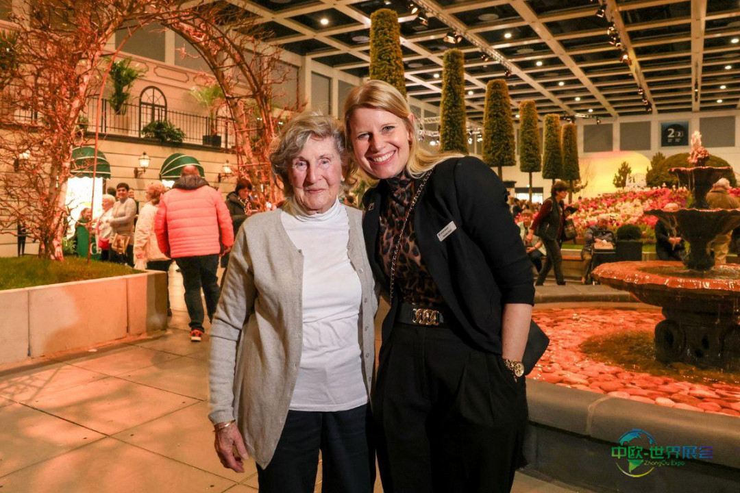 2026年柏林国际绿色周花卉厅中,101岁的英格堡·韦格纳与部门经理丹妮拉·斯塔克并肩而立,背景是高耸的锥形植物与喷泉。