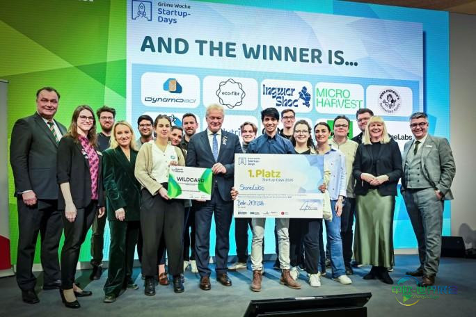 2026年创业日大赛体验农场展区舞台上,一群人手持冠军大奖支票,背景板展示着决赛企业的标识。