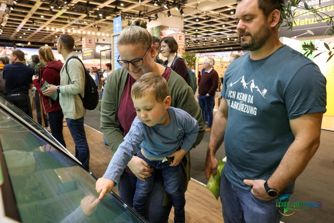 柏林绿色周展馆内，一位父亲与孩子正在指点互动触摸屏，周围是熙攘的观展人群