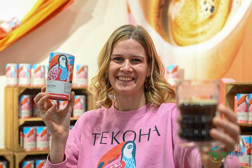 莱奥妮·特塞诺一手持有机马黛咖啡粉罐，另一手持咖啡杯，站在摆满罐装产品的货架前。